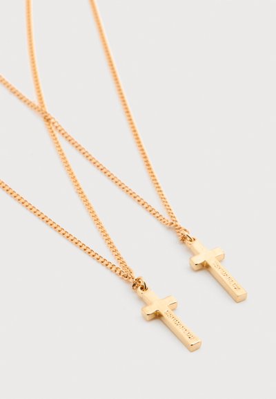 Deux pendentifs croix en or avec texte gravé pendent sur de fines chaînes en or sur un fond blanc uni.