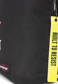 Svart tygryggsäck med en texturerad yta, med en gul etikett med "BUILT TO RESIST" och Eastpak-logotyp.