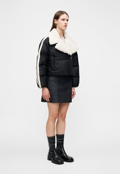 Γυναίκα ντυμένη με μαύρο μπουφάν puffer με λευκό γιακά από shearling, μαύρη μίνι φούστα με μπροστινές τσέπες και μαύρες μπότες μέχρι τη μέση της γάμπας με λευκό ράψιμο.
