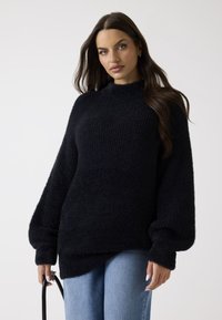 Pull en maille noire avec un col montant, des manches oversize et une texture douce, associé à un jean en denim bleu clair et un sac noir.