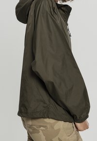 Olivgrön, lättviktsnylonjacka med huva, dragkedja i kragen, elastiska ärmslut och avslappnad passform, visad från en sidovinkel.