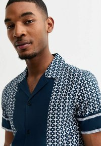 Short-sleeve shirt featuring a dark navy base with a white geometric pattern on one side, collar, and button-down front. Soft fabric texture.