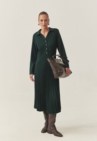 Largo vestido verde con cuello abotonado, falda plisada. Bolso de cuero marrón, botas marrones hasta la rodilla. Textura suave y corte ajustado.