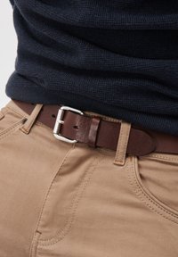 Rjav usnjen pas s srebrno kovinsko sponko, nošen preko svetlo rjavih tkaninskih hlač. Tkanina ima gladko teksturo s vidnimi šivi.