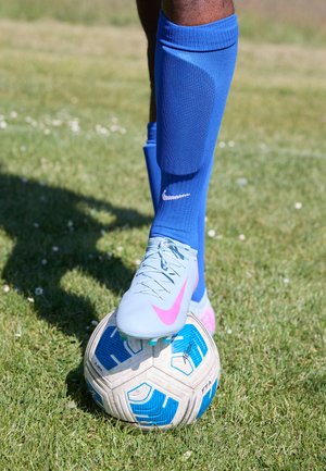 Blaue Fußballsocken, hellblaue und rosa Fußballschuhe, Füße auf einem gemusterten Ball, grüne Grasunterlage. Fokus auf Details der Schuhe und des Balls.