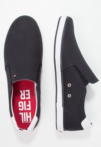 Svarta canvas slip-on skor med vita gummisulor, röd foder och metallöglor för ventilation. Har en logotyp p å innersulan.