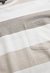 Raidallinen puuvillapaita beige ja valkoinen, jossa on rintatasku. Kangas on pehmeä, tekstuuri viimeistely horizontalilla raidoilla.