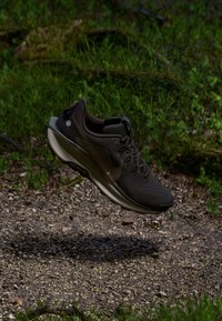 Une chaussure de sport marron foncé avec une tige en tricot texturé, un système de laçage et une semelle de couleur claire. Présente un logo sur le côté et un accent réfléchissant.