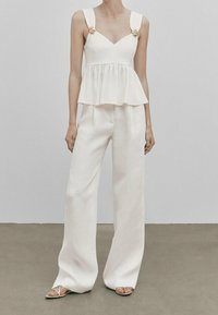 Biały top z peplum i szerokimi ramiączkami ozdobionymi motywami kwiatowymi, zestawiony z białymi, luźnymi spodniami z wysokim stanem oraz sandałami na paski.
