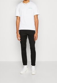 Bílý bavlněný tričko s tyrkysovou nášivkou, doplněné černými slim-fit kalhotami a bílými teniskami. Má krátké rukávy a volný střih.