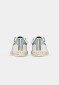 Hvite sneakers med sølvdetaljer på hælen, teksturert gummisåle og hevet skrift på mellomsålen. Glatt materialefinish.