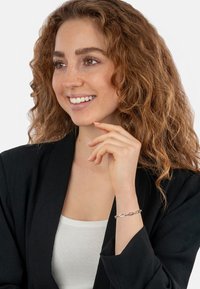 Silver chain bracelet with oval links, worn on the wrist. Model is in a black blazer over a white top, with curly brown hair.