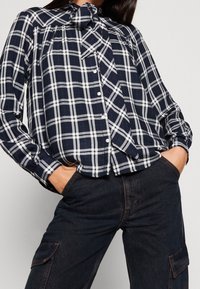 Navy blue and white plaid blouse with a bow tie collar, long sleeves, button front, and gathered detailing at the shoulders.