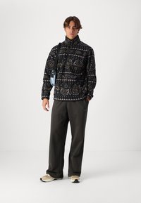 Schwarzer gemusterter Fleece-Pullover mit hohem Kragen, mit einem Paisley-Design in Grau und Beige, kombiniert mit lockeren dunklen Hosen und Sneakers.