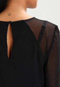 Primo piano della parte superiore della schiena di una persona che mostra un top nero con maniche trasparenti, una chiusura a bottone a forma di chiave e un piccolo tatuaggio a forma di cuore sul collo.