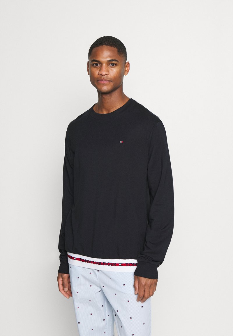 Giovane uomo che indossa una camicia nera a maniche lunghe con un piccolo logo e pantaloni azzurri chiari con un piccolo motivo ripetuto su uno sfondo neutro.