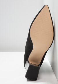 Chaussure à talon noire à bout pointu avec semelle beige et talon bloc texturé reposant contre une surface blanche.