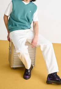 Persoon zit op een zilveren metalen koffer, draagt een turquoise mouwloos sweater, witte broek en glanzende donkerpaarse laarzen op een gele vloer.