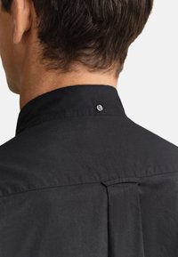 Ορατή η πλάτη και ο λαιμός άνδρα που φοράει μαύρο πουκάμισο με γιακά και κουμπί στο γιακά καθώς και κεντρική πτυχή στην πλάτη.