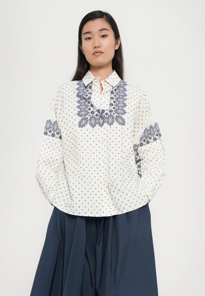 Weiße Bluse mit blauer Floralstickerei am Kragen und an den Ärmeln, mit einer Knopfleiste und verstreuten kleinen Blumenmustern.