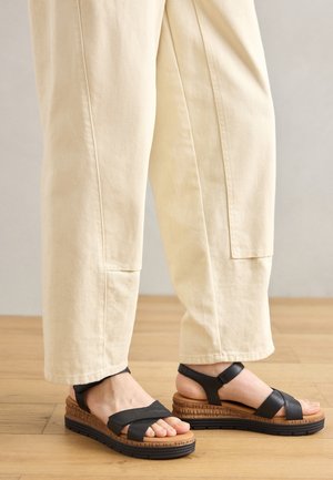 Czarne skórzane sandały na koturnie z teksturowaną brązową podeszwą i krzyżującymi się paskami, zestawione z kremowymi spodniami o szerokich nogawkach.