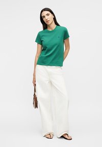 Γυναίκα με πράσινο κοντομάνικο πουκάμισο και φαρδιά λευκή παντελόνα, κρατώντας μια μικρή καφέ τσάντα, στέκεται σε λευκό φόντο.