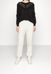 Sudadera negra con un panel de encaje y un cuello con volantes, combinada con pantalones de chándal blancos ajustados y zapatillas blancas. Diseño simple y casual.