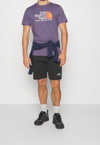 Fialové bavlněné tričko s bílým a oranžovým logem, námořnický svetr uvázaný v pase, černé šortky s logem, šedé ponožky a černé boty.