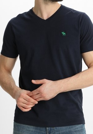 Marineblaues V-Ausschnitt-T-Shirt aus Baumwolle mit einem kleinen grünen Elch-Logo auf der Brust. Kurze Ärmel und ein lässiger Schnitt sind sichtbar.