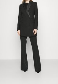 Kvinna klädd i en svart dubbelknäppt kavaj med satinrevers och matchande vida byxor, stående mot en enkel ljus bakgrund.