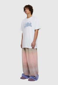 Prevelika bijela majica s plavim grafičkim dizajnom, uparena s hlačama širokog kroja u pastelnim gradijentima ružičaste i ljubičaste boje, s mekanom teksturom.