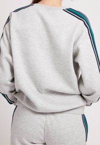 Sudadera gris hecha de un material suave, con cuello redondo, mangas largas y rayas en azul marino y verde azulado a lo largo de los hombros.
