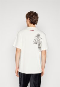 Camiseta de algodón blanca con un gráfico en la parte posterior que representa árboles y un paisaje, presenta el logotipo "HUGO" en rojo en el cuello. Mangas cortas, corte relajado.