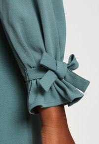 Sinisenvihreä puseron hiha, jossa on kerätty, rypytetty helma ja sidottu rusetti, valmistettu pehmeästä, sileäpintaisesta kankaasta.