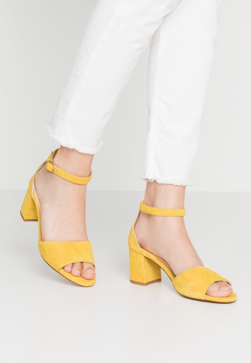 Pieds portant des sandales à talons blocs jaunes en daim avec bride à la cheville et bouts ouverts, associées à un pantalon court blanc à ourlet effiloché, sur un sol clair.