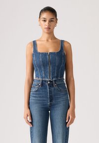 Νεαρή γυναίκα με τα μαλλιά τραβηγμένα προς τα πίσω φοράει μπλε τζιν crop top με φερμουάρ μπροστά και ψηλόμεσα μπλε τζιν, στέκεται μπροστά σε ουδέτερο φόντο.