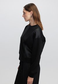 Schwarzer Pullover mit einem strukturierten Satin-Einsatz. Er verfügt über einen Rundhalsausschnitt, überschnittene Schultern und einen elastischen Saum. Glattes Material mit einer strukturierten Passform.