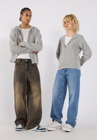 To modeller står side om side. Den ene har på seg en lys grå zip-up hettegenser, hvit t-skjorte og mørk baggy jeans. Den andre har på seg en grå hettegenser og blå jeans.