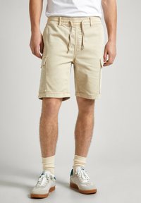 Bermudas de carga beige con cinturilla elástica, bolsillos laterales y dobladillos enrollados, hechas de una mezcla de algodón. Calzado: zapatillas beige.
