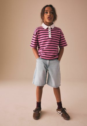 Copil în picioare cu mâinile în buzunare, purtând un tricou polo cu dungi roz și negre, pantaloni scurți din denim deschis, șosete închise la culoare și adidași maro.