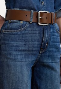 Brun lærbelte med en rektangulær sølvspenne, brukt over mørkeblå denimjeans med et klassisk fem-lomme-design.
