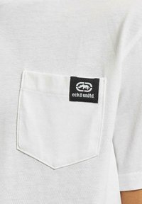 Hvid bomulds t-shirt med en brystlomme, der har et sort label med et logo. Korte ærmer og en afslappet pasform. Blød tekstur.