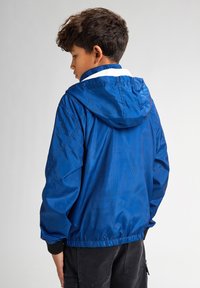 Jachetă albastră cu un model text tonal subtil, glugă, manșete elastice și bandă la talie. Materialul este fin și ușor.