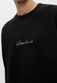Czarna bluza z miękkiej bawełny, z okrągłym dekoltem i ściągaczami, z białym haftem "United Garments" na piersi.