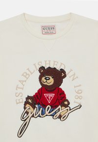 Creme genser med broderi av en brun teddybjørn som har på seg en rød genser med en trekantet logo, teksten "ETABLERT I 1981" over.