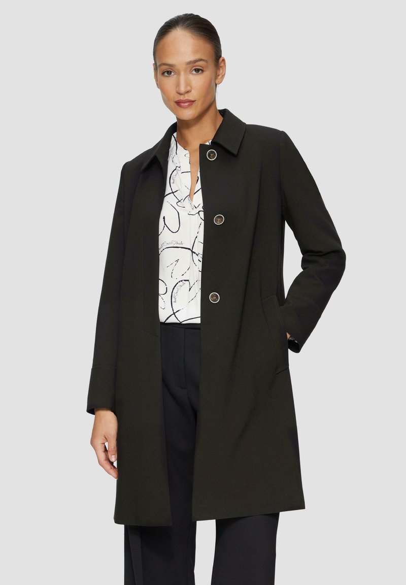 Zwarte, lange jas met een slim fit, knoopsluiting en zijzakken. Heeft een afgeschuind kraag en een gladde textuur. Gdragen over een geblokt shirt.