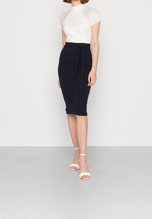 Nainen valkoisessa pitsisestä lyhythihaisessa paidassa, joka on tungettu vyötäröä korostavaan mustaan kynähameeseen, jossa on edessä solmittava yksityiskohta, yhdistettynä valkoisiin nilkkaremmisandaaleihin.