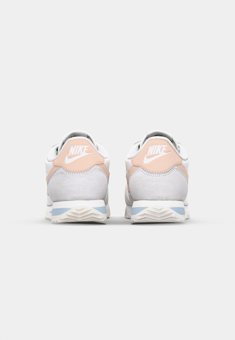 Baskets Nike avec une tige en cuir blanc, des accents en daim gris clair, un talon rose pâle et une semelle en caoutchouc blanc texturé avec des détails bleus.