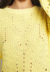 Pull en maille jaune avec un design lâche et texturé, comportant des motifs ajourés et une encolure large. Détails de coutures visibles sur l'ensemble.