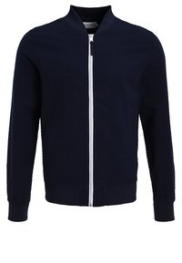 Veste zippée bleu marine en tissu texturé, avec poignets et ourlet côtelés, et une fermeture éclair blanche à l'avant, sans logos ni motifs visibles.